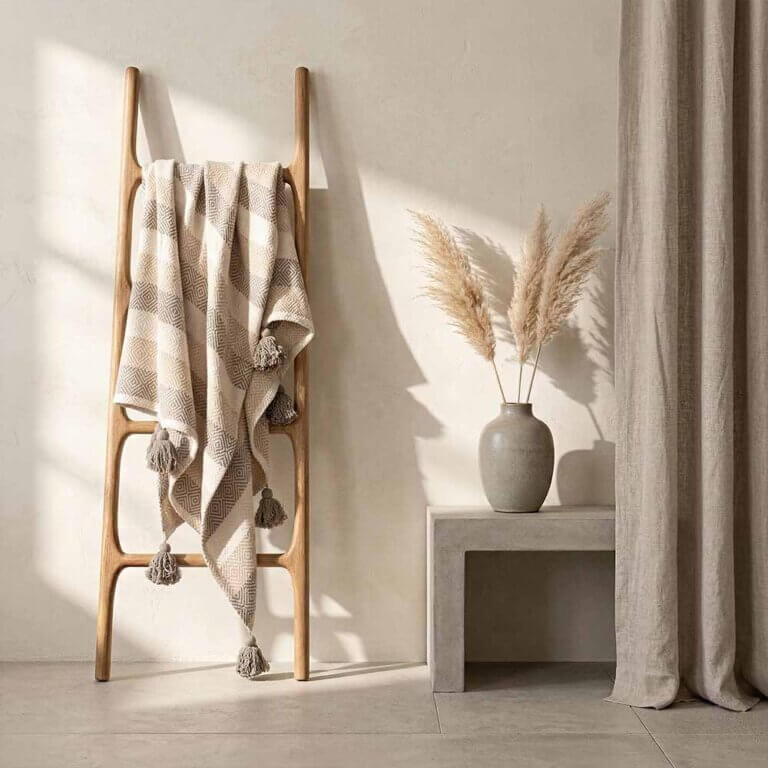 Thoughtfully Sourced, Responsibly Made: Home Decor Reimagined 9 oversized cotton throw blanket. A neutral khaki cotton woven blanket with fringe hanging on a light wood decorative ladder. The sunlight from a nearby window highlights the rhythmic, diamond-weave pattern in the stripes.