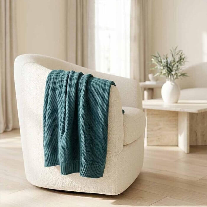 Teal cotton throw blanket in seed stitch knit draped on cream armchair in soft elegant living room with white marble fireplace