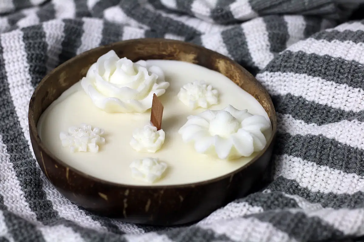 Coconut Shell Candle Dough Bowl 4 Soy candle in a coconut bowl with decorative white wax flowers and a wooden wick, presented on a gray and white knitted fabric.