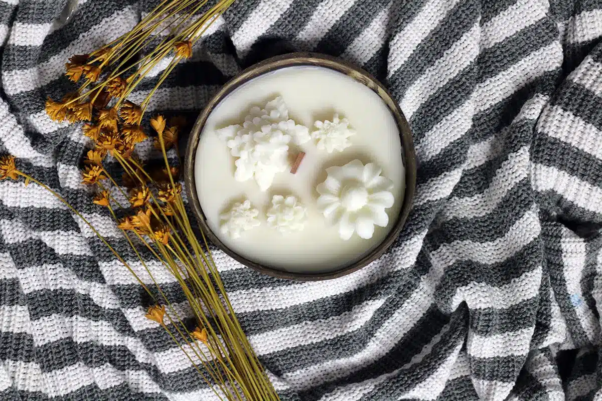 Coconut Shell Candle Dough Bowl 3 coconut bowl candles. Soy candle in a coconut bowl with decorative white flowers and a single wooden wick, placed on a gray and white knitted fabric.
