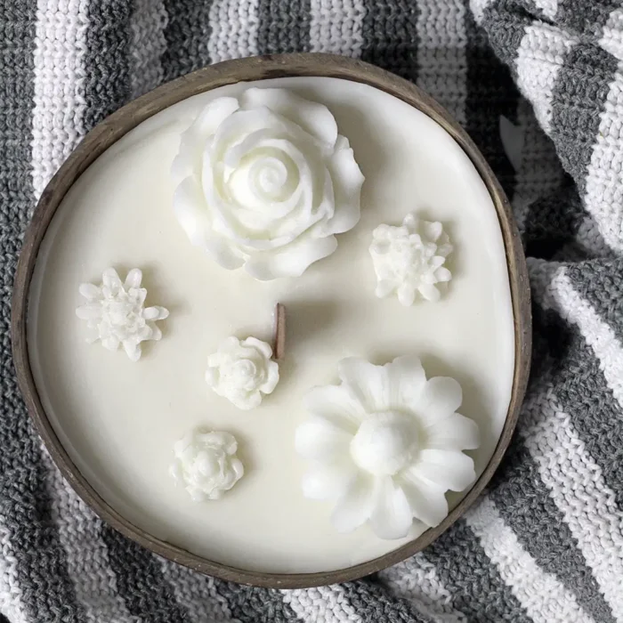 soy scented candles. Soy candle in a coconut bowl with decorative white flowers and a single wooden wick, placed on a grey and white knitted fabric.