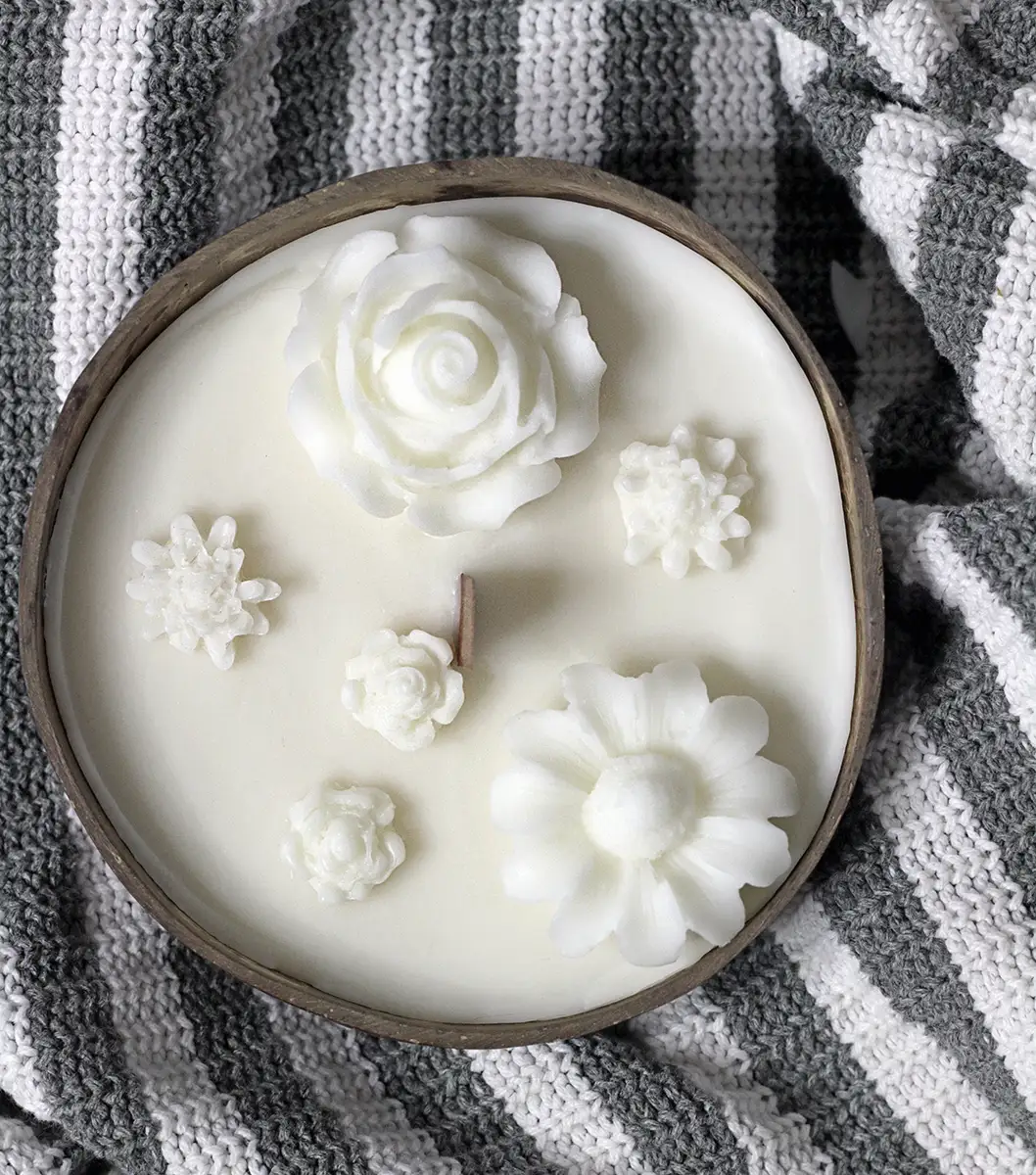 Coconut Shell Candle Dough Bowl 1 soy scented candles. Soy candle in a coconut bowl with decorative white flowers and a single wooden wick, placed on a grey and white knitted fabric.
