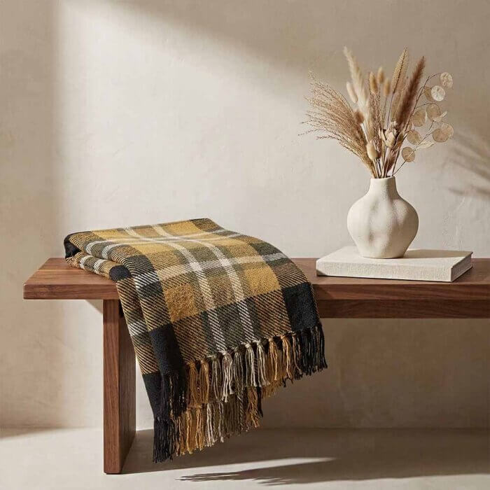 A yellow plaid blanket with long tassels draped over a rustic wooden bench. A vase with dried pampas grass stands next to the folded corner, showing the traditional tartan pattern. Yellow and Black Plaid Throw Blanket with Fringe. fall throw blanket