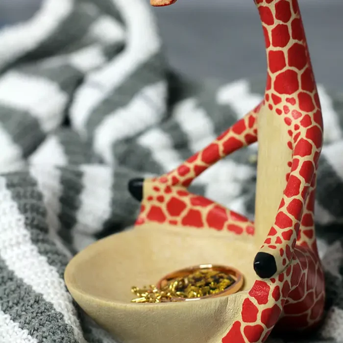 giraffe figurine Handmade giraffe shaped jewelry holder, crafted from sustainable materials, featuring a red and beige patterned body with black hooves, sitting atop a grey and white woven fabric.
