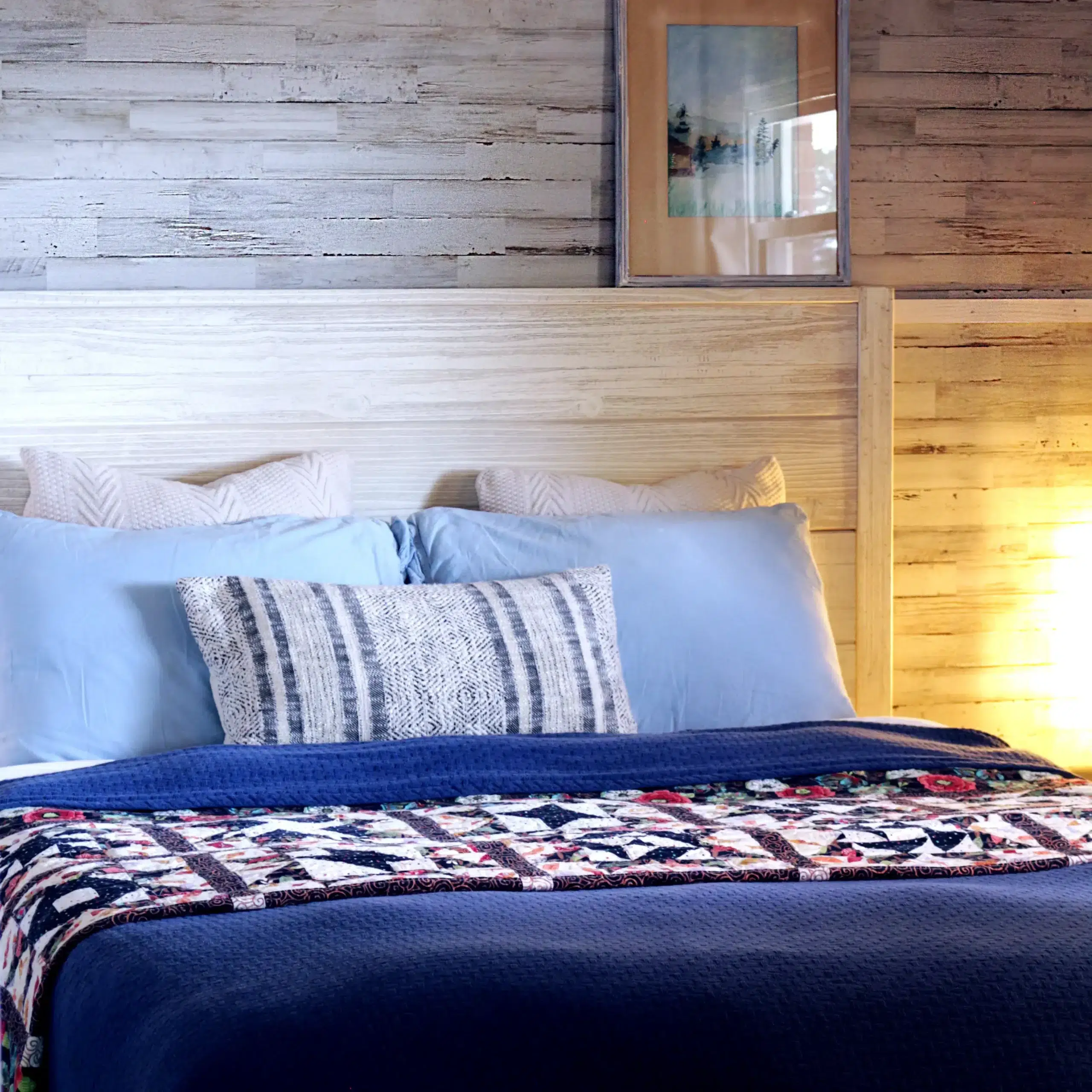 The image shows a tidy bedroom with a bed covered in a blue comforter, assorted blue pillows, and a cream-patterned one. A colorful quilt rests at the bed's foot, and a plain wooden headboard matches the light wood wall paneling behind.
