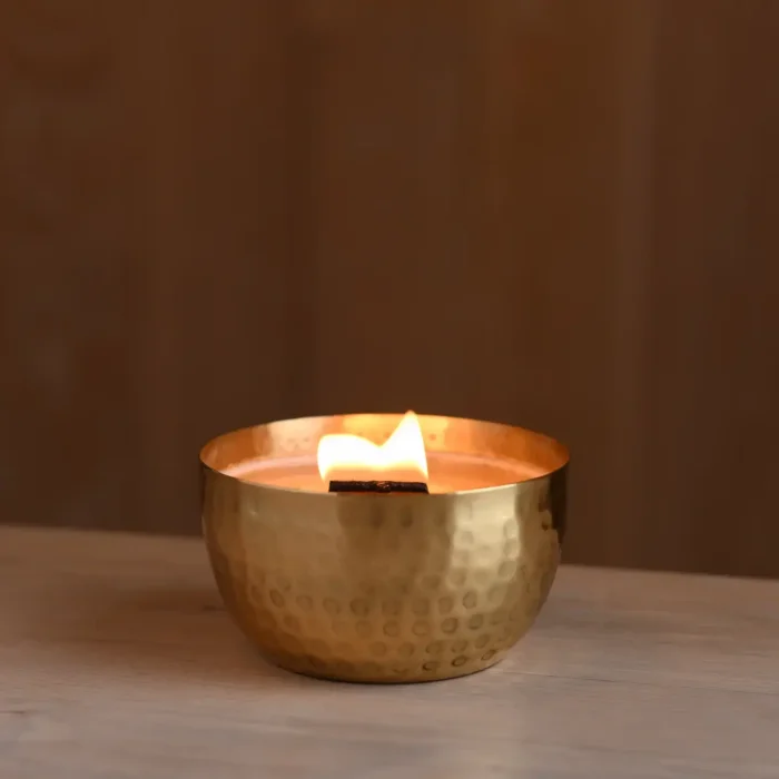 Lit Solid Brass 14oz Pure Soy Candle on Wooden Surface: A lit solid brass soy candle with a wooden wick, placed on a wooden surface with a warm, ambient background.