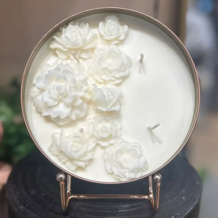 A Decorative Wax Flowers Soy Candle in a copper tin with intricate white wax flowers on top, displayed on a stand.