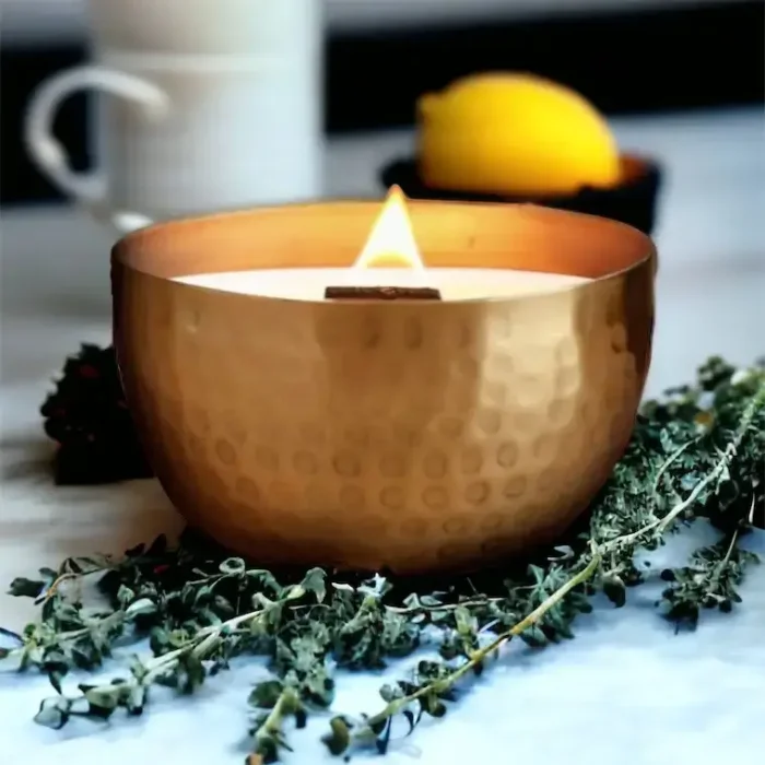 Lit Solid Brass 14oz Pure Soy Candle with Fresh Lemon and Thyme: A lit solid brass soy candle with a wooden wick, surrounded by fresh lemon and thyme, set on a kitchen countertop.