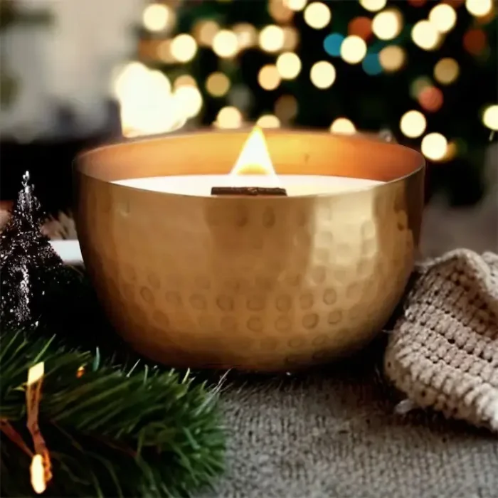 Lit Solid Brass 14oz Pure Soy Candle with Holiday Background: A lit solid brass soy candle with a wooden wick, placed in a cozy holiday setting with a decorated tree and warm lighting.
