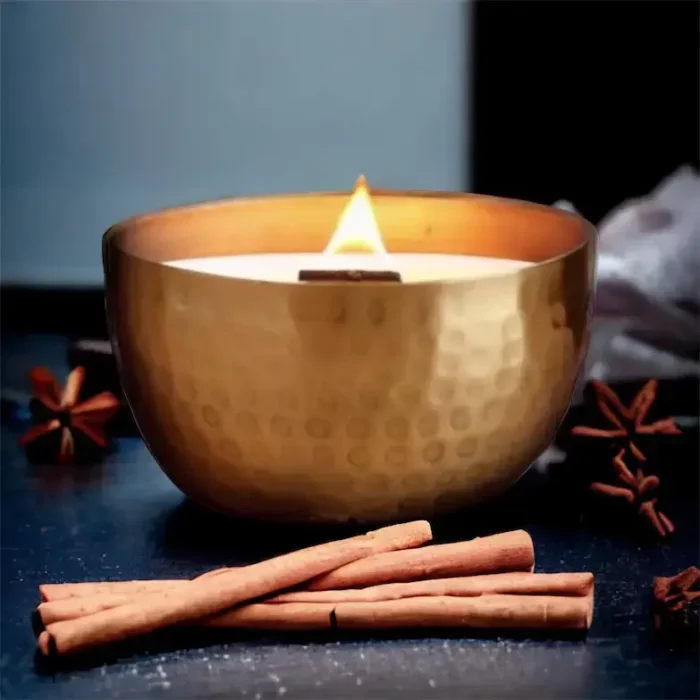 Lit Solid Brass 14oz Pure Soy Candle with Cinnamon Sticks: A lit solid brass soy candle with a wooden wick, surrounded by cinnamon sticks and star anise.
