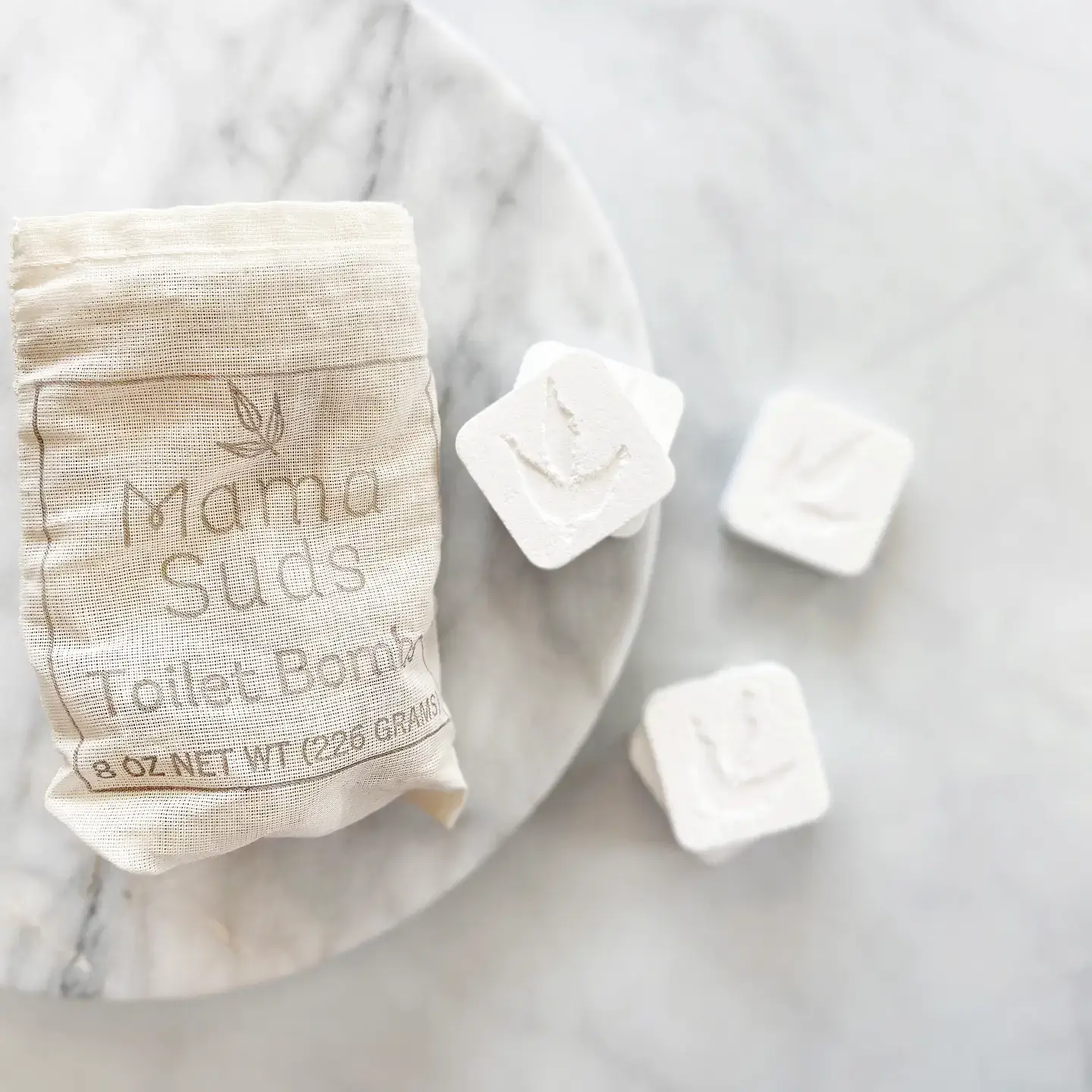 MamaSuds eco-friendly toilet cleaning bombs - 10 packs 3 MamaSuds Toilet Bomb Cleaning Tabs in a muslin bag with four individual tabs on a marble surface.