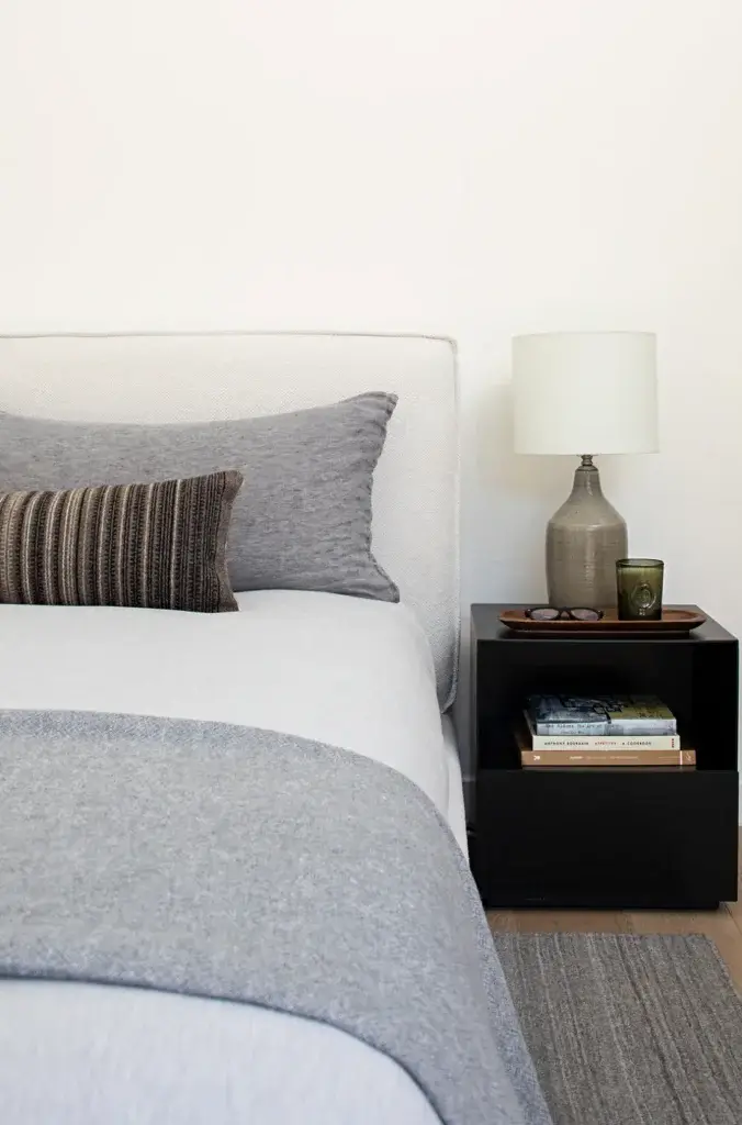 35 Nightstand Decor Ideas For a Spicy Makeover 5 Nightstand Decor Idea: A minimalist bedroom with a sleek black nightstand featuring a ceramic table lamp, a small green candle, and a wooden tray holding glasses. The shelf below holds a neat stack of books, adding a touch of personality and function. The bed is styled with neutral grey and beige tones, enhancing the calm and modern aesthetic of the space.