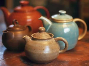 Ceramic teapots in earthy tones, including a rounded orange teapot and a light blue Asian-style teapot, sitting on a rustic wooden surface.