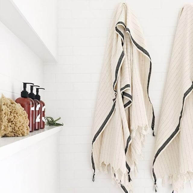 Silvia 100% Cotton Turkish Towels - Loomia 2 a bright, clean bathroom setting with a minimalist aesthetic. Two cream-colored Turkish towels with black stripes hang prominently on the right side of the frame. These towels feature a delicate fringed edge, adding a touch of texture and artisanal charm.