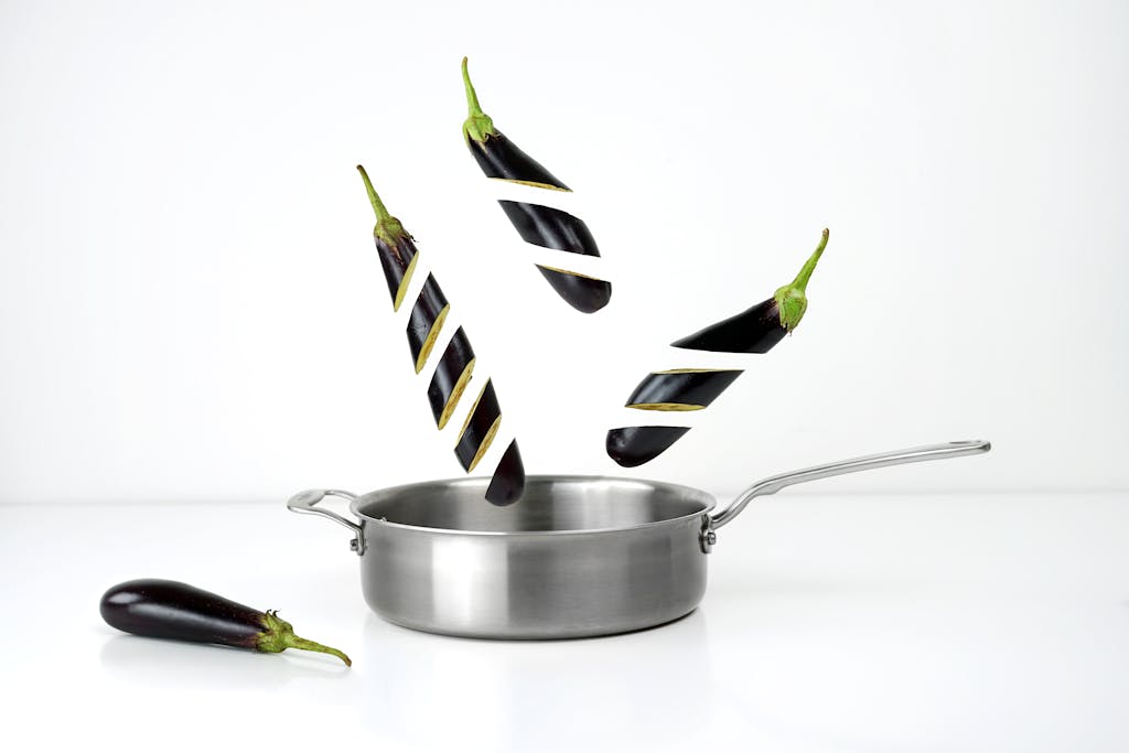 The Healthiest Material for Cookware| Your Guide to the Best Materials for Pots & Pans in 2025 9 Creative image of sliced eggplants floating above a stainless steel pan against a white background.