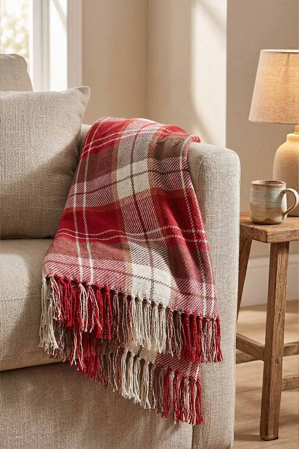 Red and White Plaid Throw Blanket with Fringe 1 A cozy red throw blanket with a red and white plaid pattern draped over a soft beige sofa. The blanket has long, hand-tied fringe tassels hanging down. A warm lamp and a small plant in the background make the room feel inviting.
