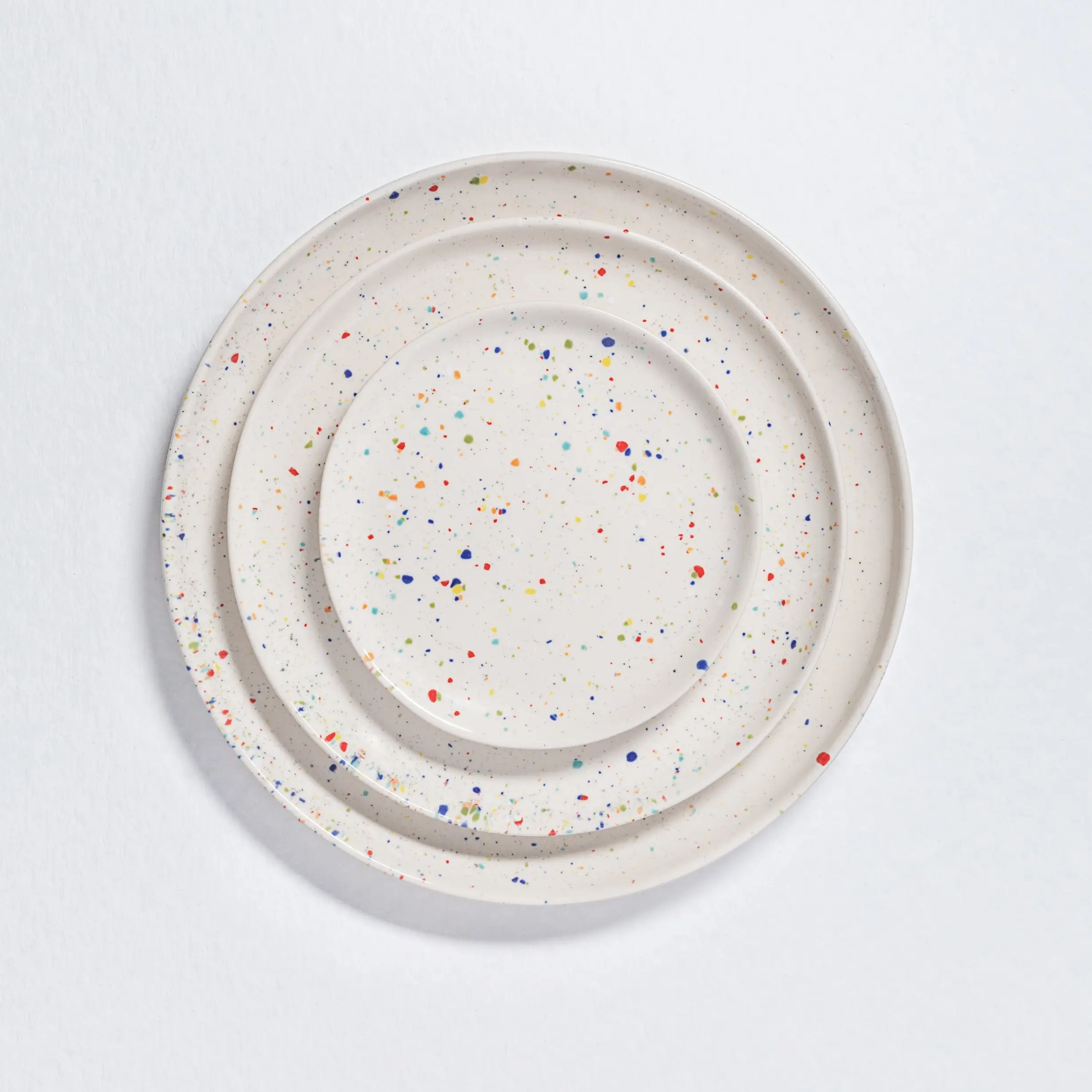 16-Piece White Ceramic Dishes Set | Made In Portugal 2 Top-down view of three round white ceramic plates in different sizes. The organic, wavy edges show the handmade Portuguese craftsmanship, and colorful dots are scattered across the white surface. colorful dishe sets