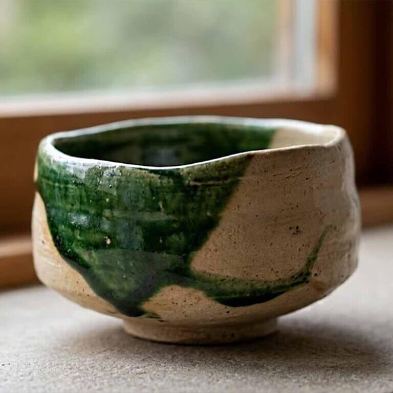 What Is a Chawan? A Guide to the Japanese Matcha Bowl 6 A traditional Japanese Oribe chawan tea bowl, hand-shaped with an intentionally irregular and playful form, vivid deep green copper glaze flowing across part of the surface, contrasting with raw beige clay