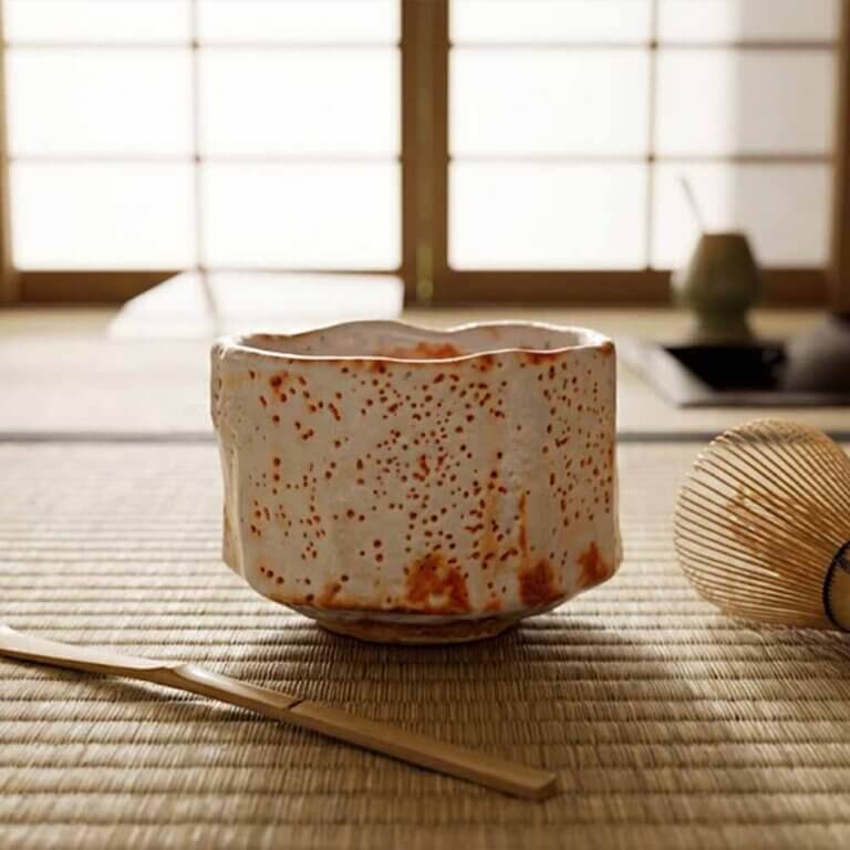 What Is a Chawan? A Guide to the Japanese Matcha Bowl 5 A handcrafted Shino chawan tea bowl used in a traditional Japanese tea ceremony, thick milky white glaze with warm orange fire marks, soft rounded handmade shape, placed on tatami mat with bamboo whisk and tea scoop