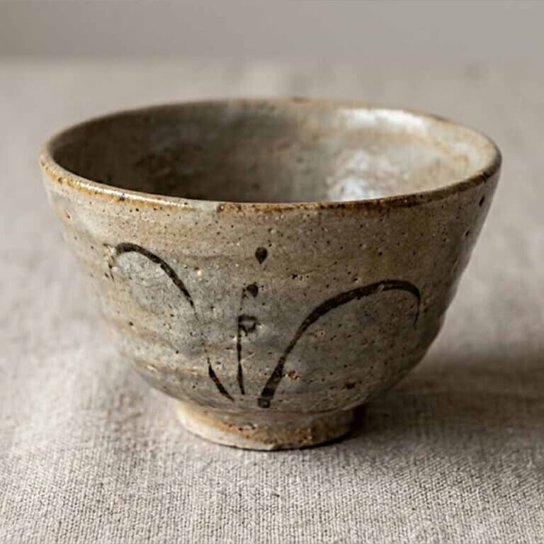 What Is a Chawan? A Guide to the Japanese Matcha Bowl 4 A traditional Japanese Karatsu chawan tea bowl from Saga Prefecture, hand-shaped with a slightly uneven, organic form, earthy clay body with rough texture, natural ash glaze in muted tones of beige, gray, and brown, simple hand-painted brushstroke design
